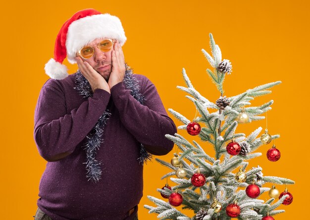 오렌지 배경에 고립 된 카메라를보고 얼굴에 손을 유지 장식 된 크리스마스 트리 근처에 서 안경으로 목 주위에 산타 모자와 반짝이 갈 랜드를 입고 혼란 중년 남자