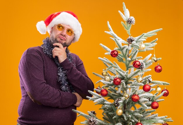 산타 모자와 장식 된 크리스마스 트리 근처에 서있는 안경으로 목 주위에 반짝이 화환을 입고 혼란 중년 남자