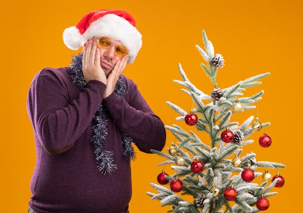 Смущенный мужчина средних лет в новогодней шапке и мишурной гирлянде на шее в очках стоит возле украшенной елки