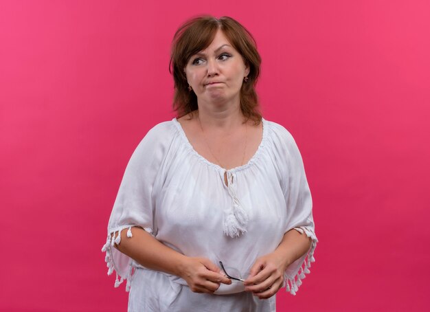 Confused middle-aged woman holding glasses on isolated pink wall