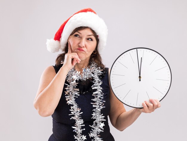 Confused middle-aged woman wearing santa hat and tinsel garland around neck holding clock keeping hand on chin looking up isolated on white background