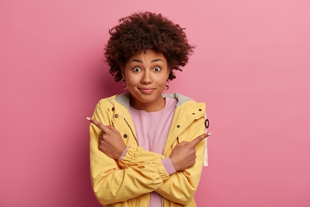 Confused positive woman with Afro hair points both index fingers in different sides, demonstrates two directions, shows something left and right