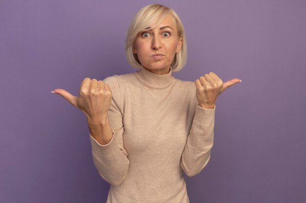 Confused pretty blonde slavic woman points at sides on purple