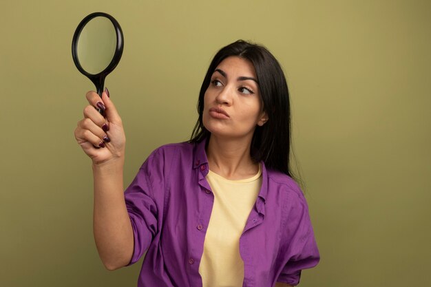 Confused pretty brunette woman holds and looks at magnifying glass isolated on olive green wall