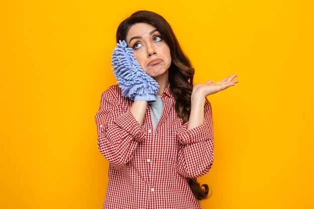 Confused pretty caucasian cleaner woman holding microfiber cleaning glove close to her head and keeping hand open