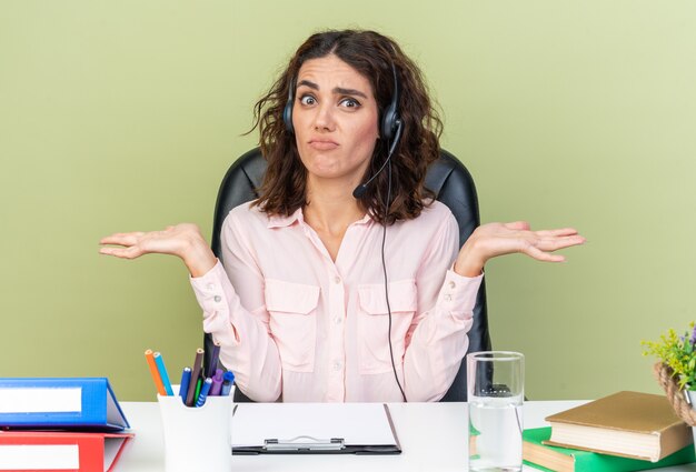 Confused pretty caucasian female call center operator on headphones sitting at desk with office tools keeping hands open isolated on green wall