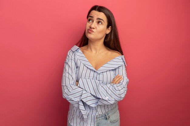 Confused pretty caucasian woman stands with crossed arms looking at side on pink