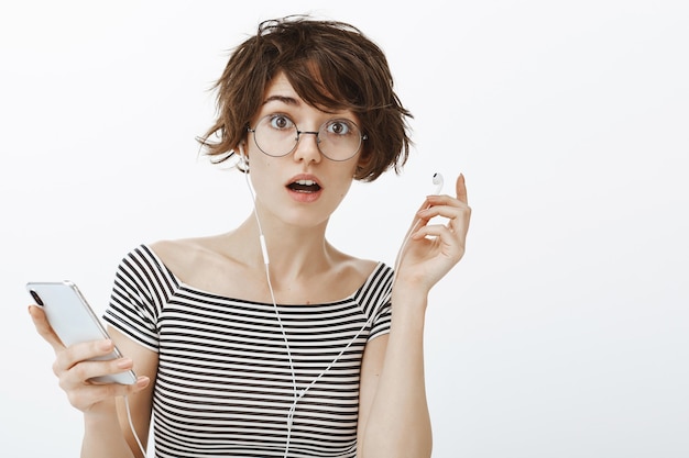 Free photo confused pretty woman in glasses take-off earphone to hear something, holding smartphone