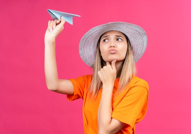 A confused pretty young woman in an orange t-shirt wearing sunhat thinking with hand on chin flying paper airplane on a pink wall