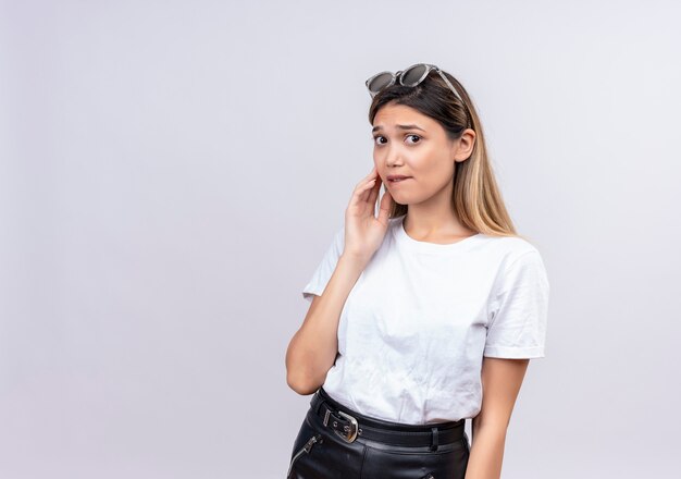 A confused pretty young woman in white t-shirt wearing sunglasses on her head keeping hand on her cheek while looking on a white wall