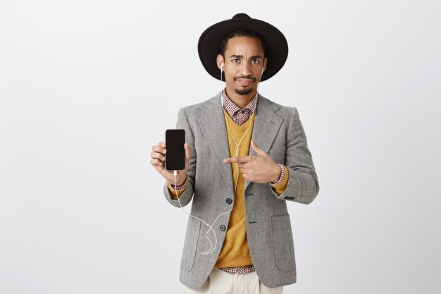 Confused and puzzled african-american guy in suit pointing finger at mobile phone with skeptical expression