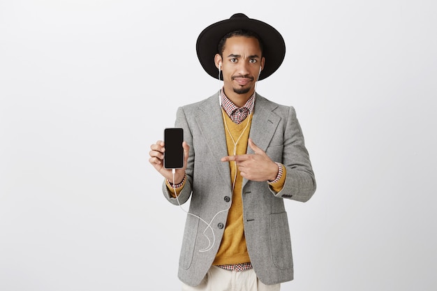 Смущенный и озадаченный афро-американский парень в костюме, скептически показывающий пальцем на мобильный телефон