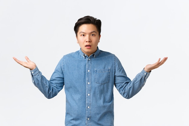 Confused and puzzled asian man in shirt cant understand what happening, raising hands sideways and shrugging, waiting explanation, being wondered, standing questioned white background