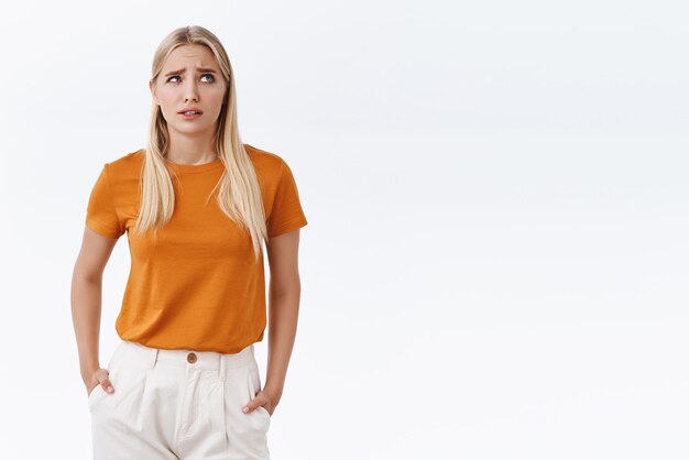 Free photo confused puzzled cute clueless blond girl in orange tshirt hold hands in pockets furrow eyebrows and look up pondering something thinking unsure standing doubtful over white background