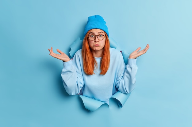 Confused puzzled red haired young questioned female shrugs shoulders and spreads palms sideways stands clueless in ripped hole of blue paper hole