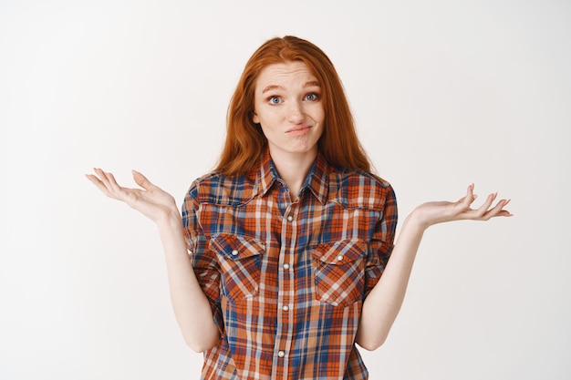 Confused redhead girl dont know anything, shrugging and smirking clueless, standing unaware against white wall