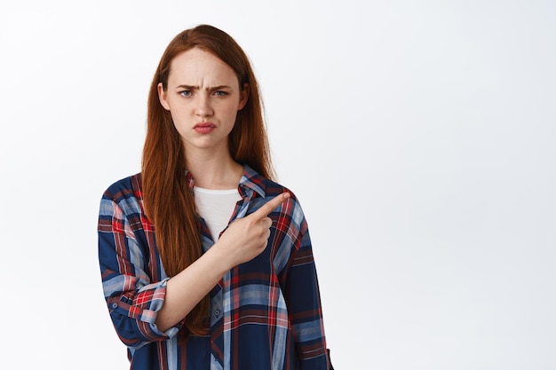 Confused redhead girl frowns suspicious, pointing at upper right corner with displeased face expression, showing something strange, standing over white background