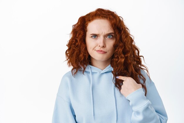 Confused redhead girl pointing at herself with frowning face being suspicious hear something strange about her white background