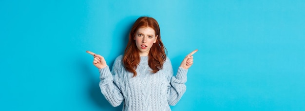 Free photo confused redhead girl in sweater pointing fingers sideways staring at camera doubtful standing again