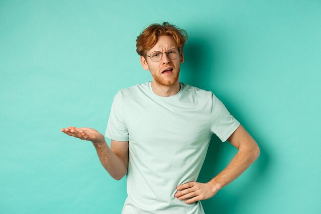 Confused redhead man trying to understand something, squinting clueless and stretch out one hand, looking puzzled, standing over mint background