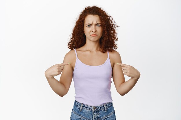 困惑した赤毛の女性は眉をひそめ、自分自身に指を向け、気分を害したり非難されたり、白い背景に対してタンクトップに立っています。