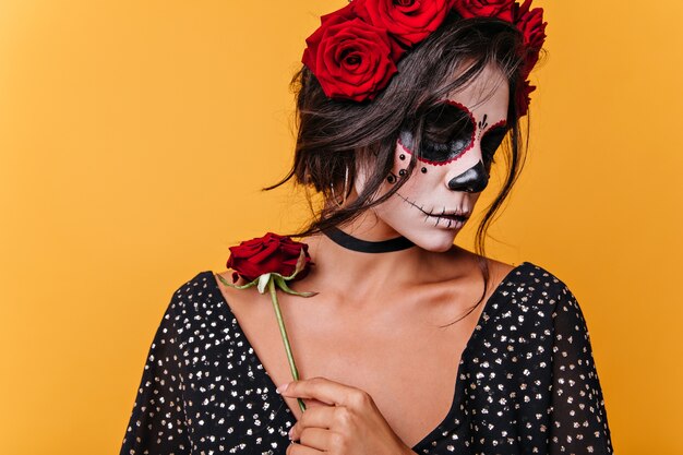 Confused romantic girl with face art in form of skull holds rose on collarbone. Brunette posing in black top on orange wall.