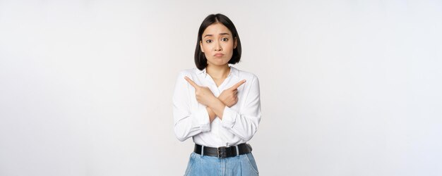 Confused sad asian girl cant decide pointing fingers left and right sulking and shrugging indecisive cannot choose standing over white background