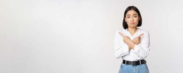 Confused sad asian girl cant decide pointing fingers left and right sulking and shrugging indecisive cannot choose standing over white background