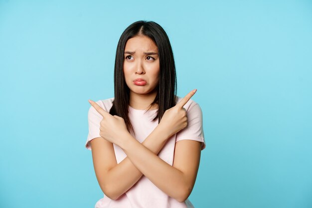 Confused and sad asian girl pointing sideways looking clueless or indecisive problem with choosing p...