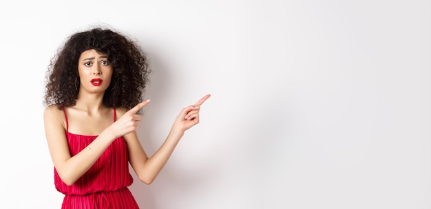 Confused and sad woman with curly hair wearing evening dress and makeup pointing fingers right and f