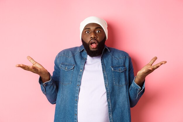 Confused and shocked african american man spread hands sideways and drop jaw, staring with awe and amazement at camera, standing over pink background clueless