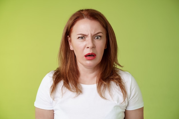 Confused shocked gasping middleaged redhead woman cringe frustrated puzzled open mouth speechless freak out strange shocking scene stand green wall perplexed disappointed