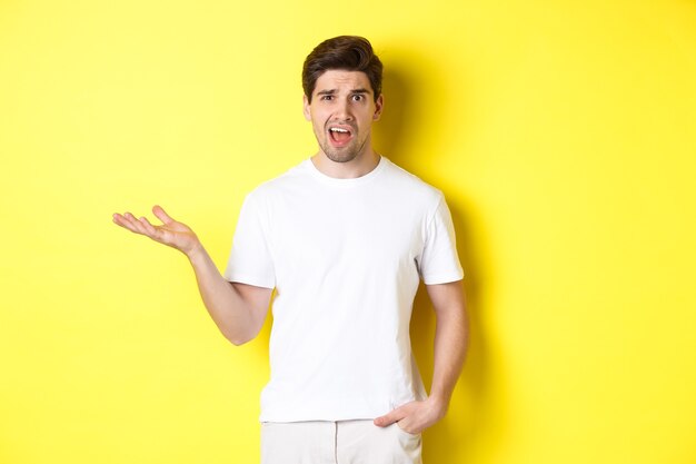 Confused and shocked man complaining, raising one hand and looking bothered, standing near yellow copy space