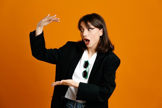 オレンジ色の背景に分離された黒いジャケットを着ている若い美しい女性のサイズを示す混乱