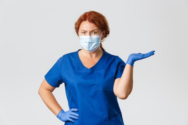 Confused skeptical female doctor, dentist in scrubs, face mask and gloves, shrugging, pointing right and frowning disappointed