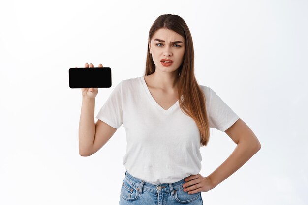 Смущенная скептически настроенная молодая женщина показывает горизонтальный пустой экран смартфона и смотрит на вопрос, оценивая что-то онлайн, делится странным веб-сайтом или сетью, стоящей на белом фоне