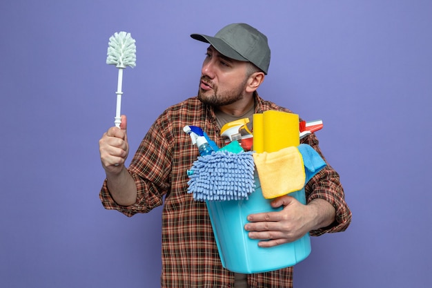 Confused slavic cleaner man holding cleaning equipment and looking at toilet brush