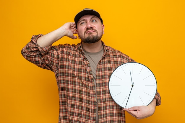 Confused slavic cleaner man holding clock and scratching his head looking at side 