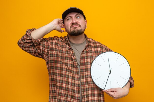 Confused slavic cleaner man holding clock and scratching his head looking at side 