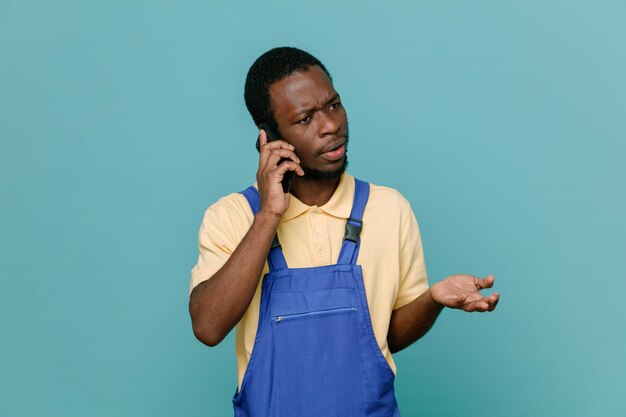 Confused speaks on the phone young africanamerican cleaner male in uniform isolated on blue background