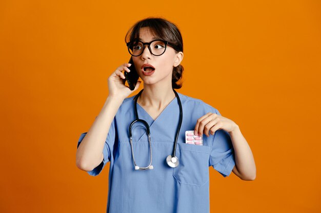 Confused speaks on the phone young female doctor wearing uniform fith stethoscope isolated on orange background