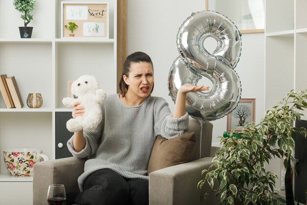 Confused spreading hand beautiful girl on happy women day holding teddy bear sitting on armchair in living room