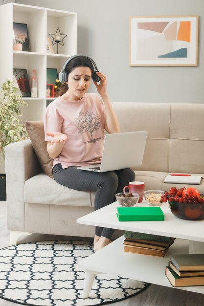 Confused spreading hand young girl with laptop wearing headphones sitting on sofa behind coffee table in living room