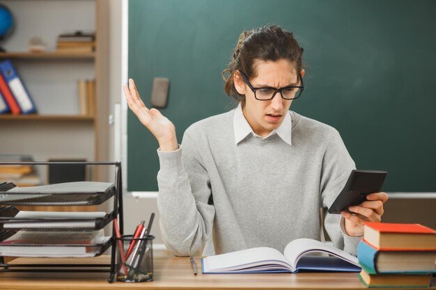 教室で学校のツールをオンにして机に座っている電卓を持っている手の若い男性教師を広げて混乱