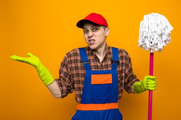 Confused spreading hands young cleaning guy wearing uniform and cap with gloves holding mop 