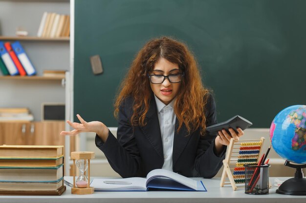 混乱した手を広げる若い女性教師が教室で学校の道具を使って机に座っている電卓を持って眼鏡をかけている