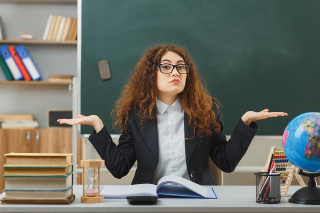 教室で学校の道具を使って机に座っている眼鏡をかけている若い女性教師が手を広げて混乱している