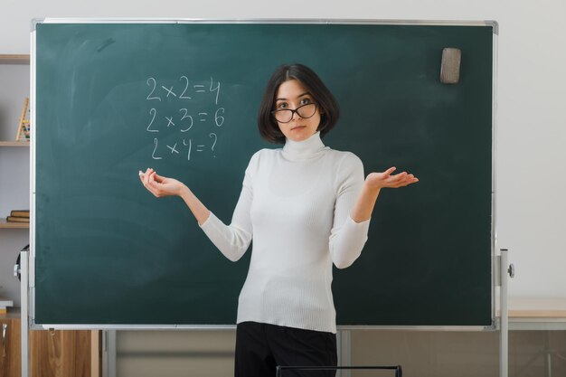 confused spreading hands young female teacher wearing glasses standing in front blackboard and writes in classroom