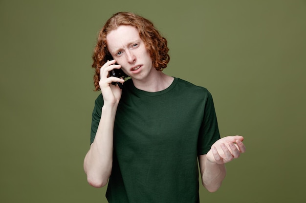 Foto gratuita mani sparse confuse il giovane bel ragazzo che indossa una maglietta verde parla al telefono isolato su sfondo verde