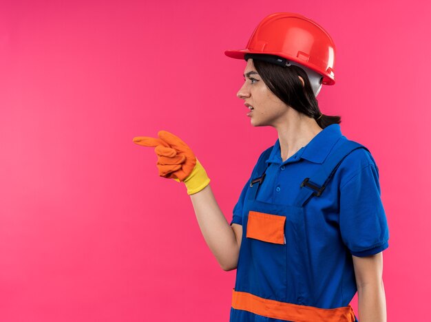 Confused standing in profile view young builder woman in uniform wearing gloves points at side 